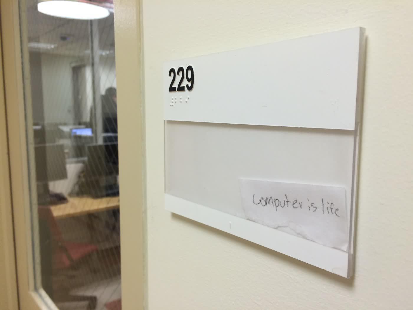 A sign with a room number outside an NYU computer lab, with a small handwritten slip of paper reading 'Computer is life'.