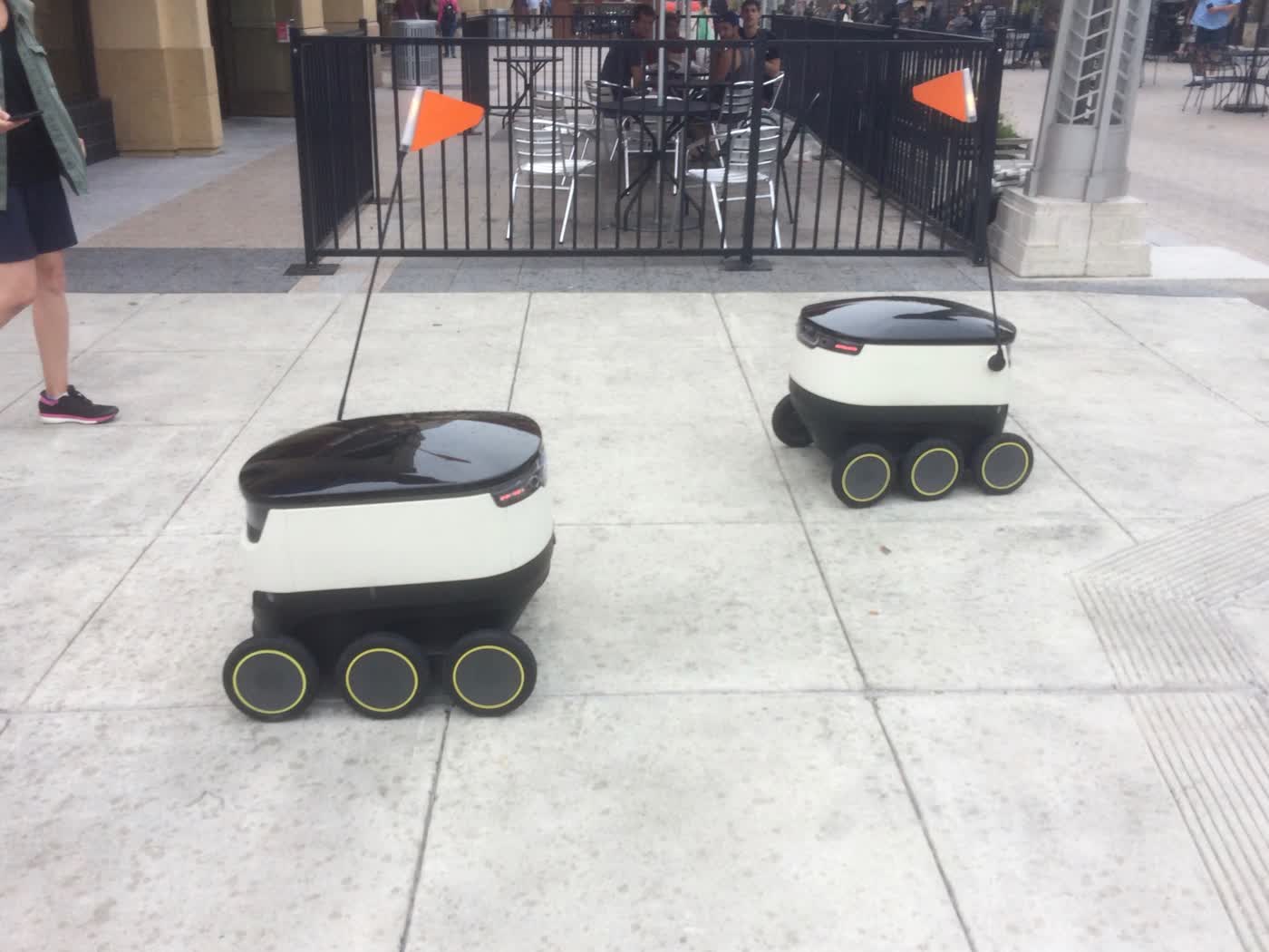 A few small boxy six-wheeled robots zipping around on a sidewalk.