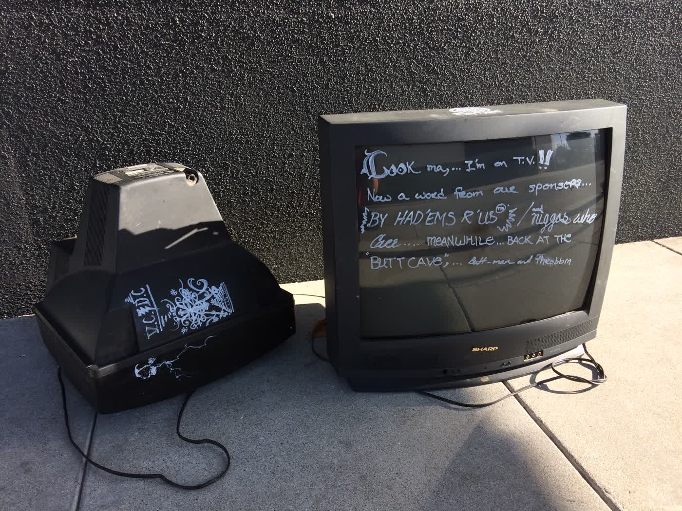 A pair of discarded CRT TVs, the left one lying face-fown, the right one standing up facing the camera. The left one has some art on its side with the initials “V.C. D.C.”. The right one has writing on the screen that says: ‘Look ma,… I’m a T.V.!! Now a word from our sponsors… BY HAD’EMS R’ US™/and n*ggas who Care… MEANWHILE… BACK AT THE “BUTT-CAVE”,… Butt-man and throbbin‘