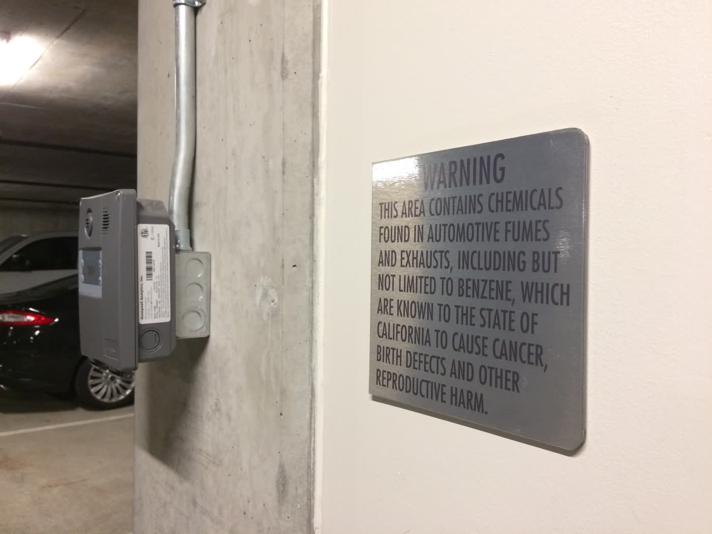 A sign in a parking lot that says “WARNING: THIS AREA CONTAINS CHEMICALS FOUND IN AUTOMOTIVE FUMES AND EXHAUSTS, INCLUDING BUT NOT LIMITED TO BENZENE, WHICH ARE KNOWN TO THE STATE OF CALIFORNIA TO CAUSE CANCER, BIRTH DEFECTS AND OTHER REPRODUCTIVE HARM.”