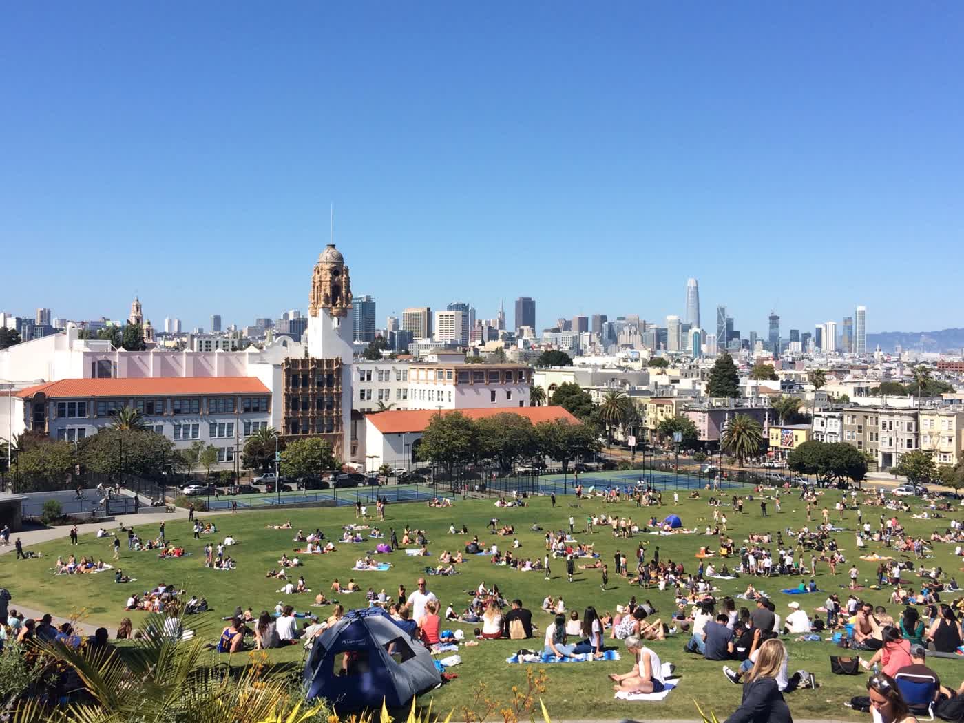 A view from the top of Dolores Park, with a huge number of sunbathers on the grass. The Basilica is visible at the far end of the park, and the towers of downtown SF can be seen far in the distance.