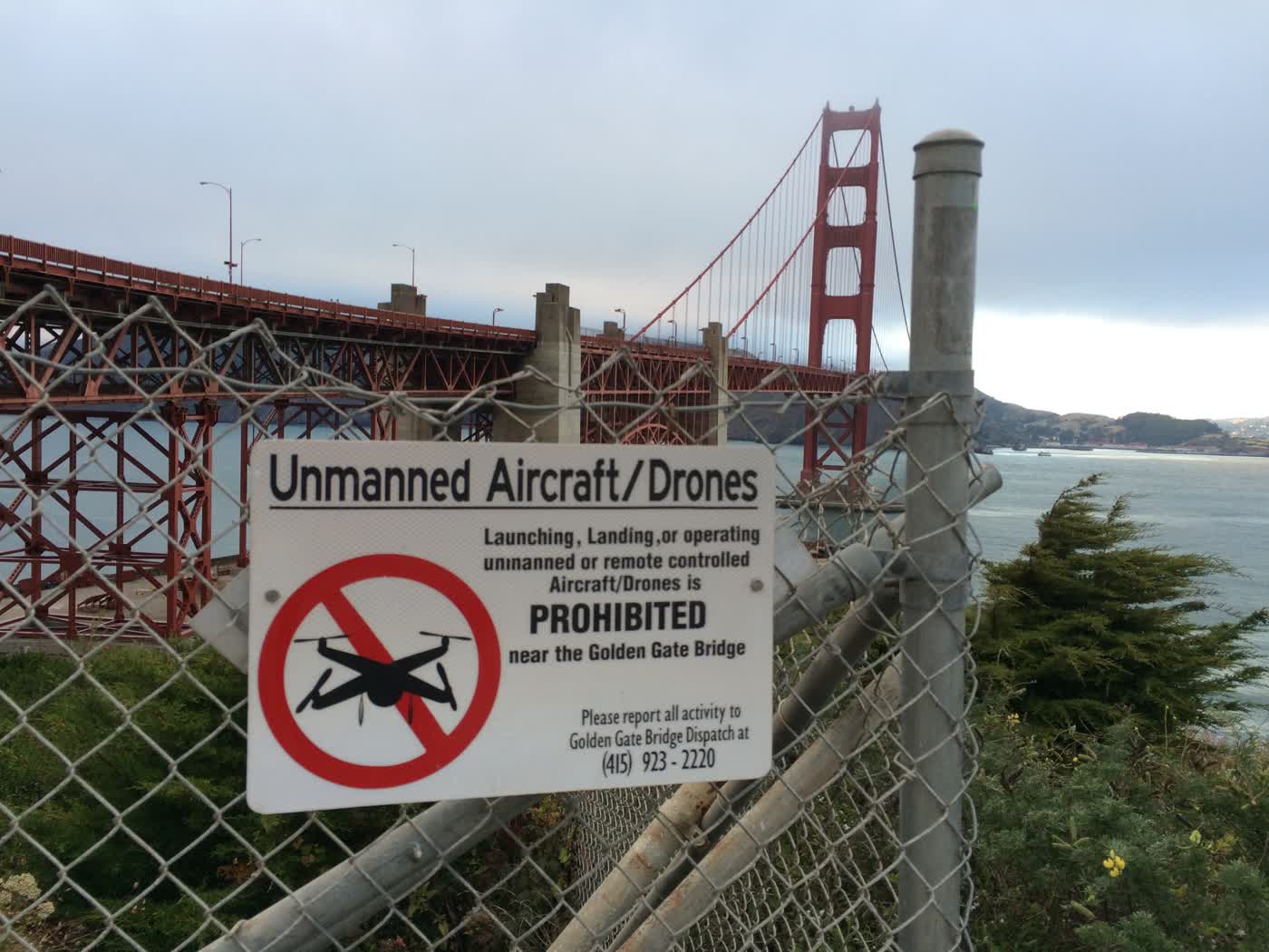 A picture with the Golden Gate Bridge in the background, but with a sign saying ”Unmanned Aircraft/Drones PROHIBITED” on the corner of a chain link fence in the foreground.