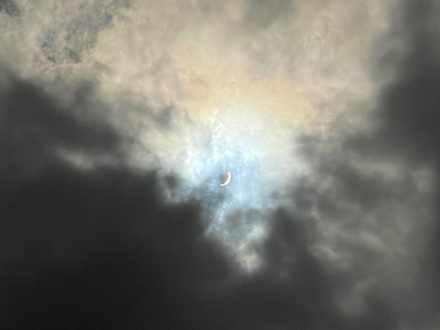 The sun just after totality, a tiny sliver visible through iridescent clouds.